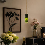 Cargar imagen en el visor de la Galería, Single green jade cylinder pendant light glowing beside framed botanical wall art and flowers